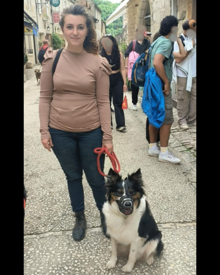 Chienne muselée entouré de plusieurs personnes. On voit un chien plus loin, c'est un lieu fréquenté. Elle semble intriguée mais calme.